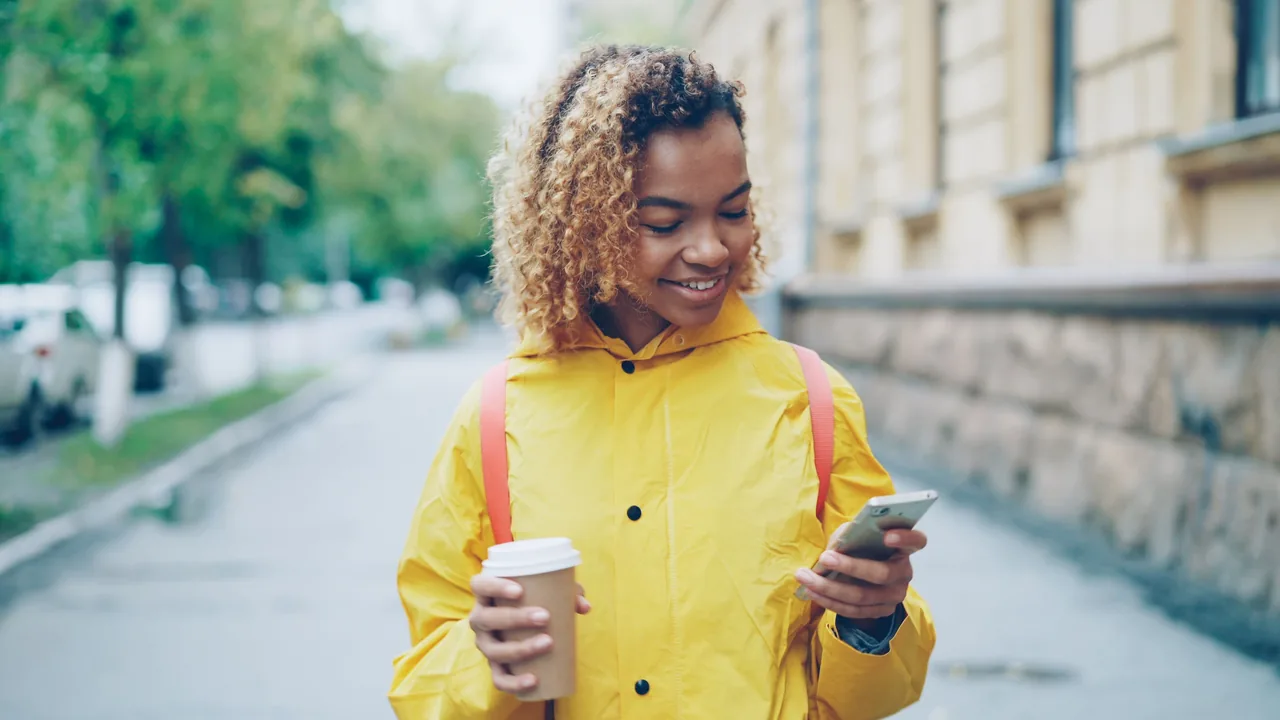 Ragazza con impermeabile giallo e caffè da asporto che sorride leggendo lo smartphone per strada
