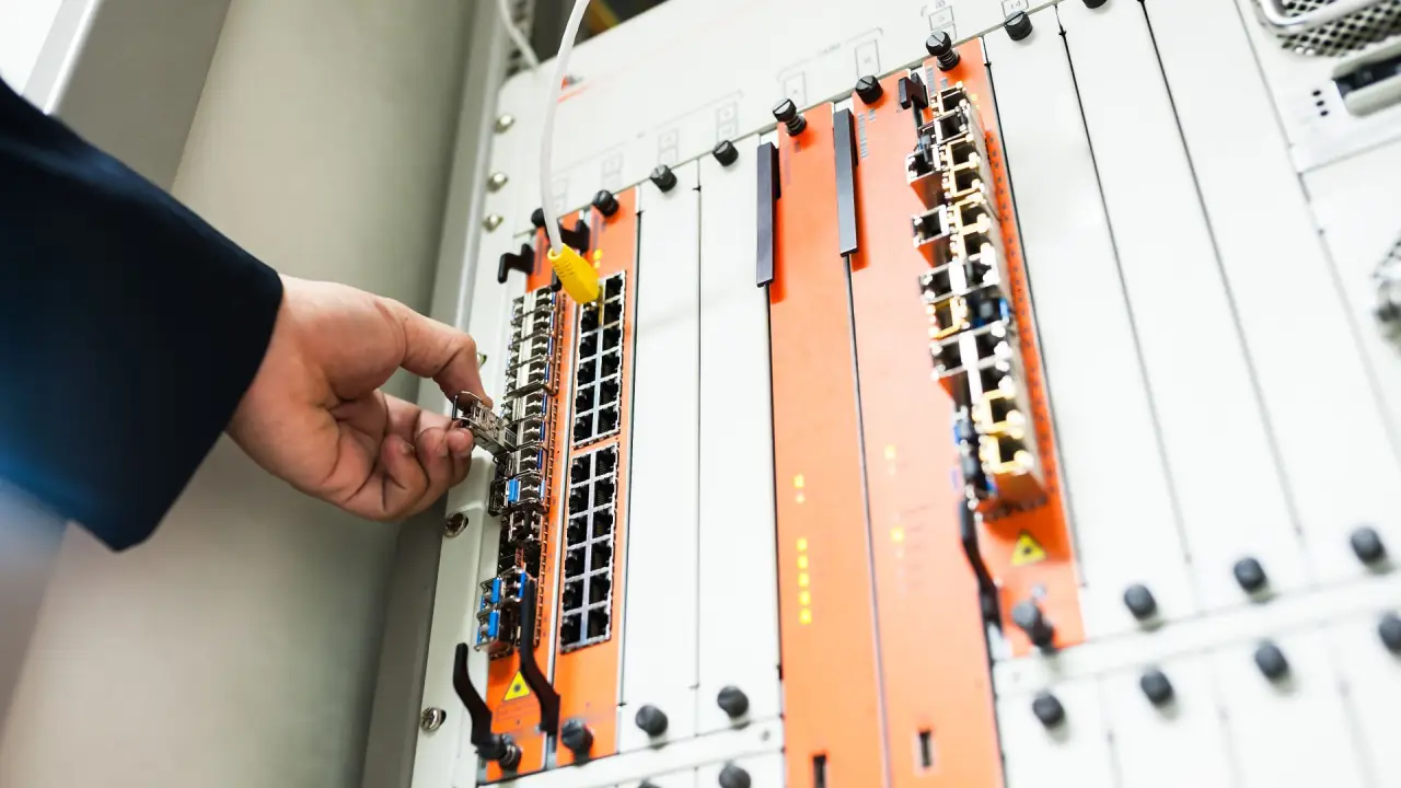 Mano che inserisce un connettore di rete nelle porte di uno switch montato in un armadio rack, con moduli arancioni e grigi