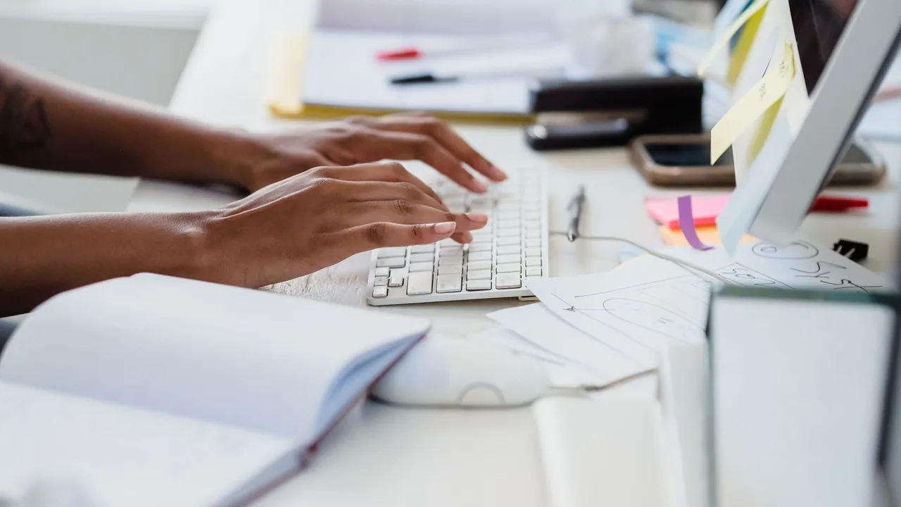 Mani che inseriscono dati alla tastiera su una scrivania d'ufficio con documenti e post-it