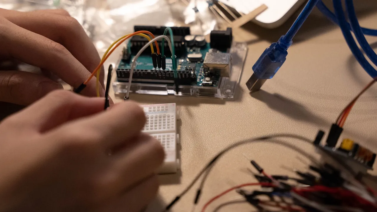 Mani che inseriscono un cavetto in una piccola breadboard bianca collegata con fili colorati a una scheda elettronica, con un cavo USB blu e un fascio di cavetti sul tavolo